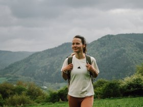 Ein Wanderer genie&szlig;t die frische Bergluft und die atemberaubende Aussicht auf die sanften H&uuml;gel und dichten W&auml;lder. Die Wolken am Himmel verleihen der Szenerie eine mystische Atmosph&auml;re, w&auml;hrend die Natur in voller Bl&uuml;te steht. Diese Idylle l&auml;dt dazu ein, die Seele baumeln zu lassen und die Sch&ouml;nheit der Umgebung zu erkunden.