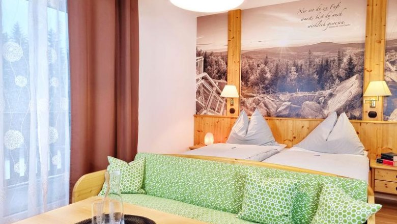 Gem&uuml;tliches Doppelzimmer mit zwei Betten, gr&uuml;nem Sofa und gro&szlig;em Wandbild einer Landschaft.