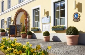 Eingang eines gelben Geb&auml;udes mit Blumen und Schildern.