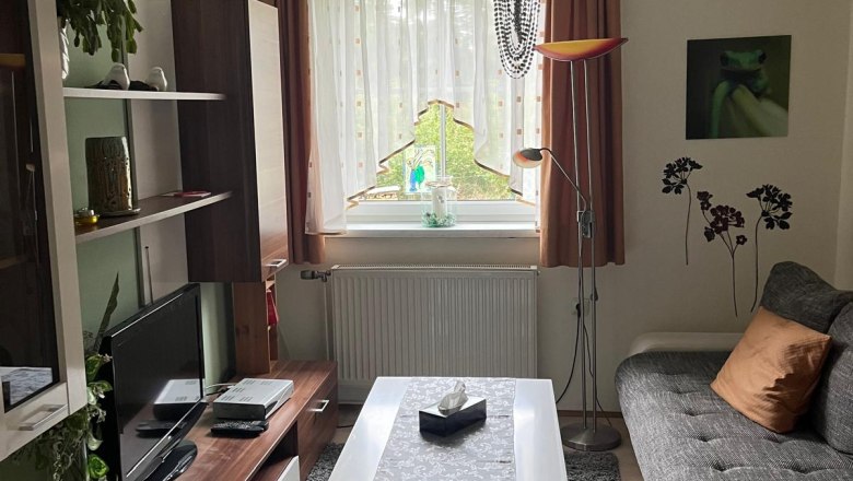 Gem&uuml;tliches Wohnzimmer mit Sofa, Couchtisch, Teppich und Fernseher. Ein Kronleuchter h&auml;ngt von der Decke, Pflanzen und Dekorationen sind sichtbar.