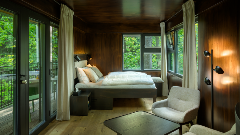 Innenansicht eines modernen Baumhauses mit Bett, Sesseln und gro&szlig;en Fenstern mit Blick ins Gr&uuml;ne.