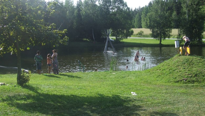 Menschen baden in einem Naturteich umgeben von B&auml;umen und Wiese.