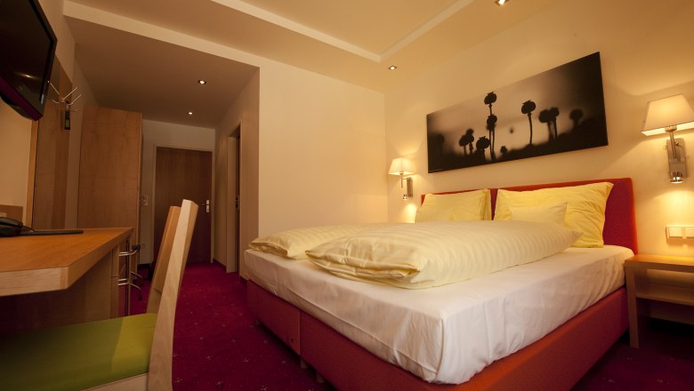Ein gem&uuml;tliches Hotelzimmer mit Doppelbett, Schreibtisch und Wandbild. Warme Beleuchtung und rote Teppiche schaffen eine einladende Atmosph&auml;re.