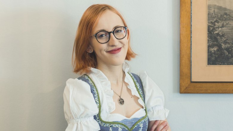 Eine Frau mit Brille und Dirndl l&auml;chelt vor einem Bild an der Wand.