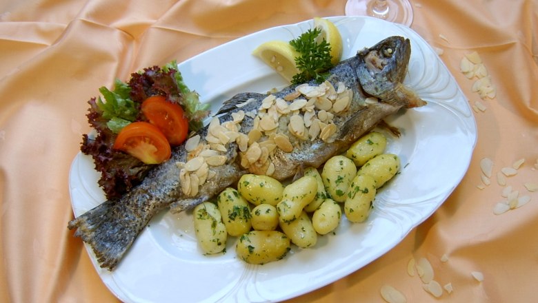 Ein Teller mit einer gebratenen Forelle, bedeckt mit Mandeln, serviert mit Kartoffeln, Salat und Zitronenscheiben auf einem orangefarbenen Tuch.