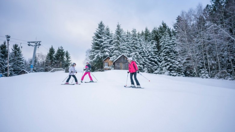 Drei Skifahrer auf einer verschneiten Piste mit B&auml;umen im Hintergrund.