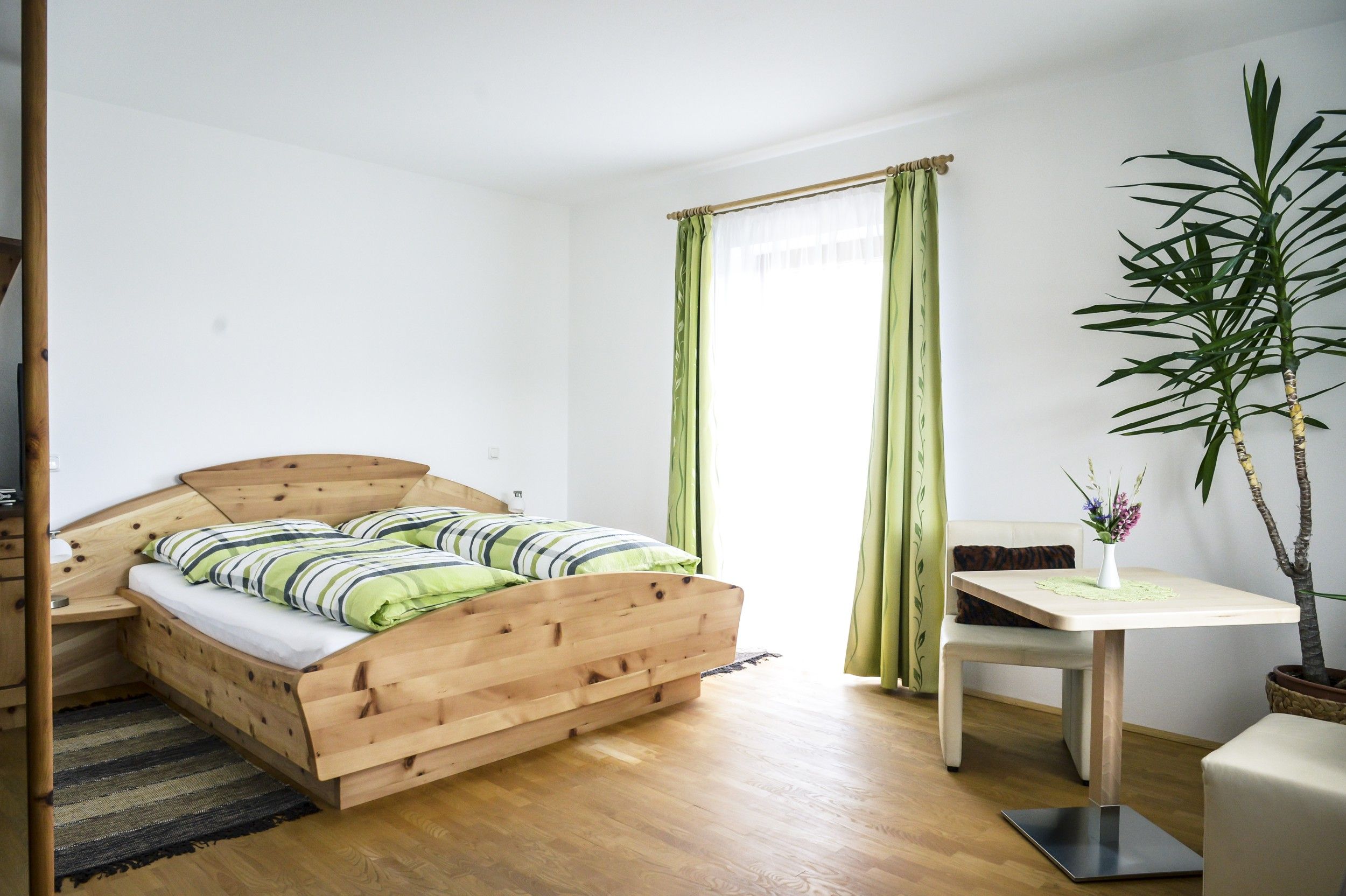 Helles Zimmer mit Holzbett, grün-weiß gestreifter Bettwäsche, Tisch mit Vase und Pflanze, grüne Vorhänge.