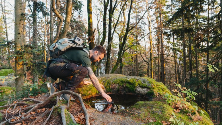 Ein Mann mit Rucksack kniet im Wald und ber&uuml;hrt Wasser in einer Felsmulde.