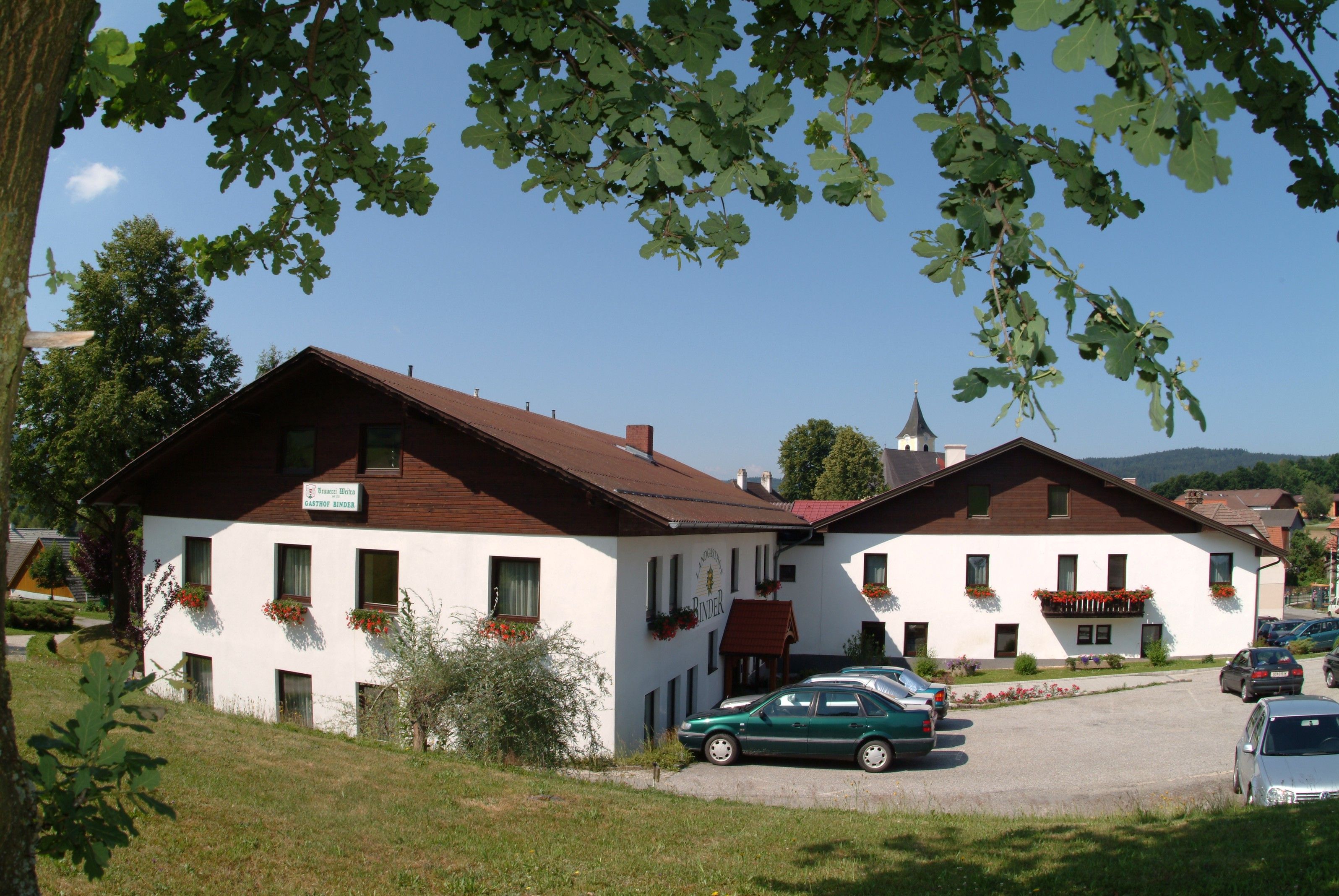 Ein Landgasthof mit weißen Wänden und braunem Dach, umgeben von Bäumen und Autos auf einem Parkplatz.