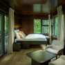 Innenansicht eines modernen Baumhauses mit Bett, Sesseln und gro&szlig;en Fenstern mit Blick ins Gr&uuml;ne.