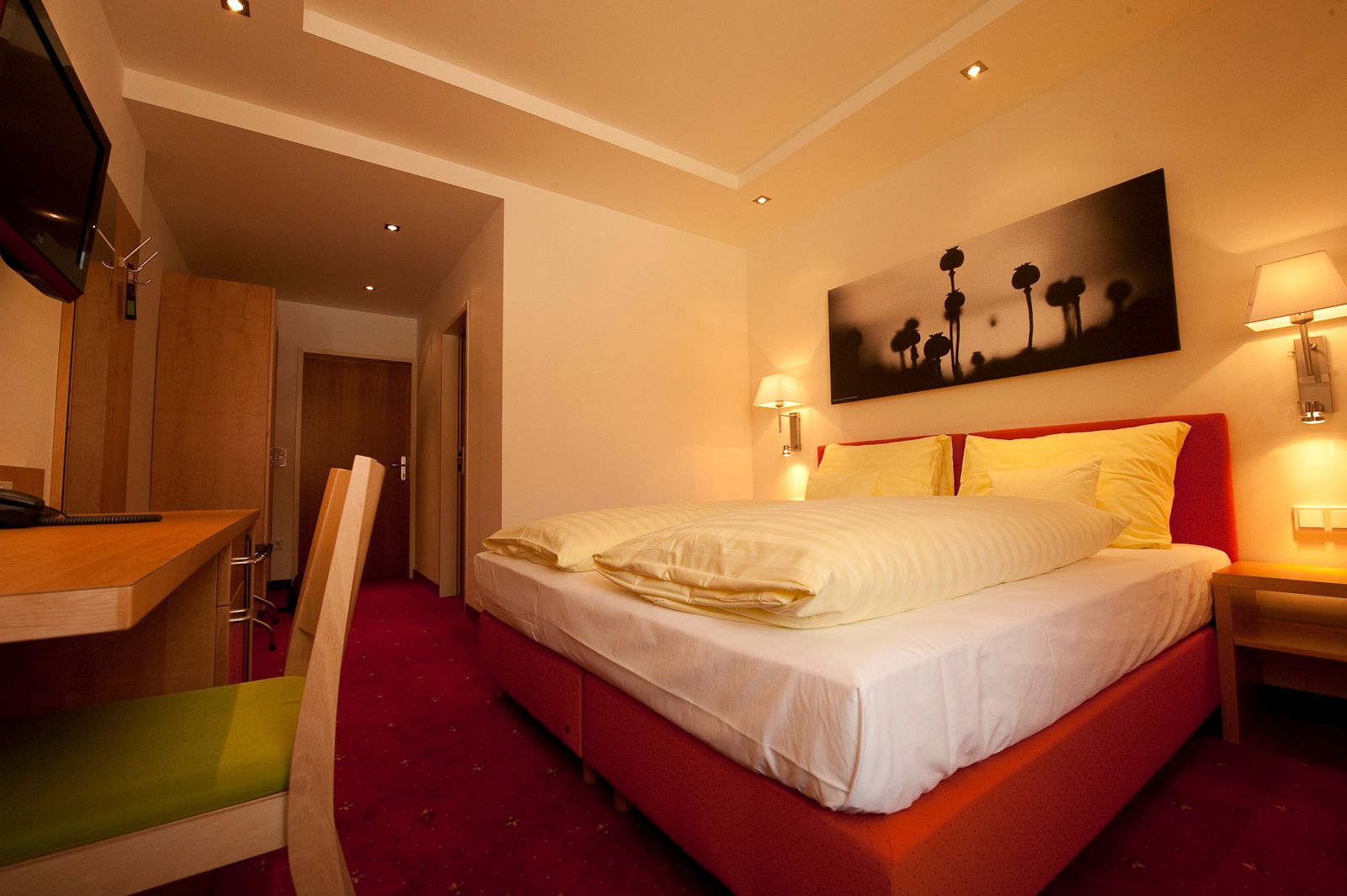Ein gemütliches Hotelzimmer mit Doppelbett, Schreibtisch und Wandbild. Warme Beleuchtung und rote Teppiche schaffen eine einladende Atmosphäre.