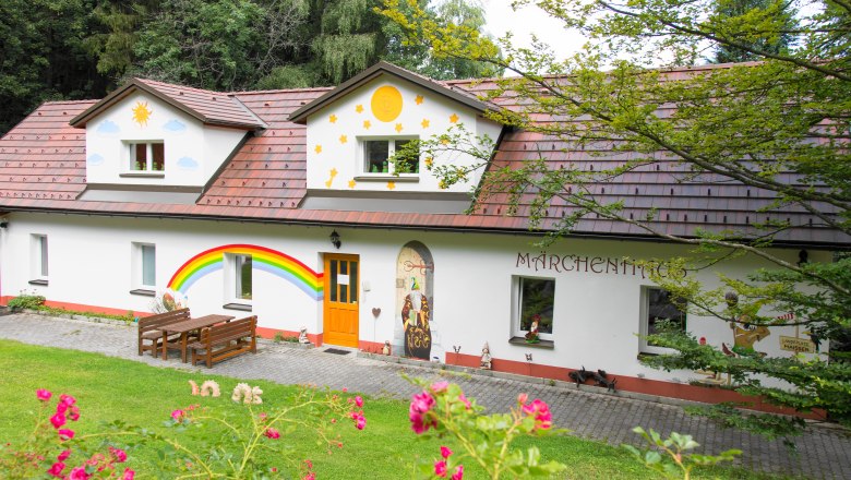 Ein bunt bemaltes Haus mit Märchenmotiven und einem Regenbogen an der Wand, umgeben von grüner Natur.