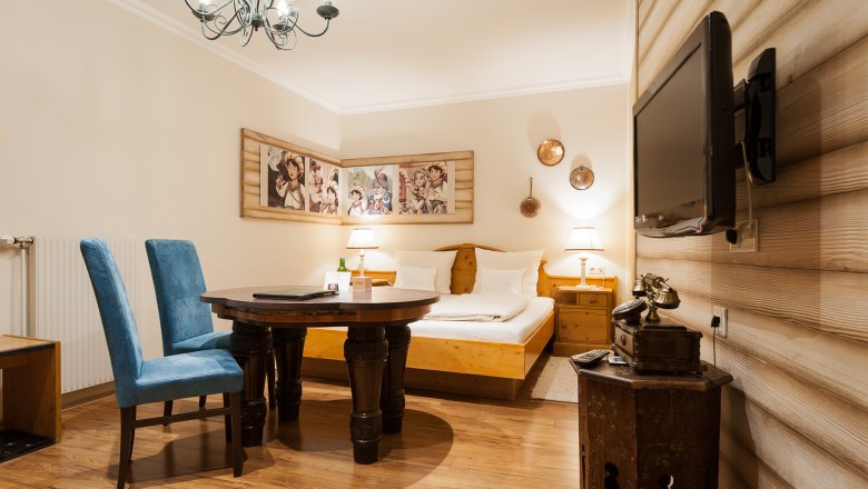 Ein gem&uuml;tliches Hotelzimmer mit Holzm&ouml;beln, einem Doppelbett, blauen St&uuml;hlen, einem runden Tisch und einem Flachbildfernseher an der Wand.