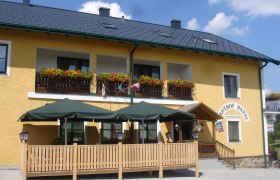 Gelbes Geb&auml;ude mit Balkon, Blumen und Terrasse mit Sonnenschirmen.