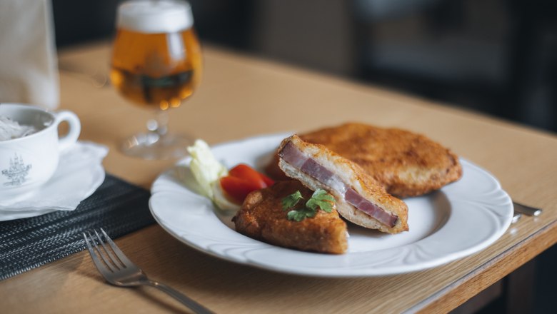 Ein Teller mit Schweinsschopf im Erd&auml;pfelmantel, serviert mit Salat und einem Glas Bier im Hintergrund.