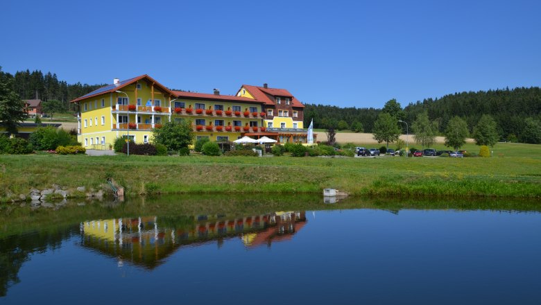 Ein gelbes Geb&auml;ude mit roten D&auml;chern spiegelt sich in einem Teich, umgeben von gr&uuml;ner Landschaft und B&auml;umen.