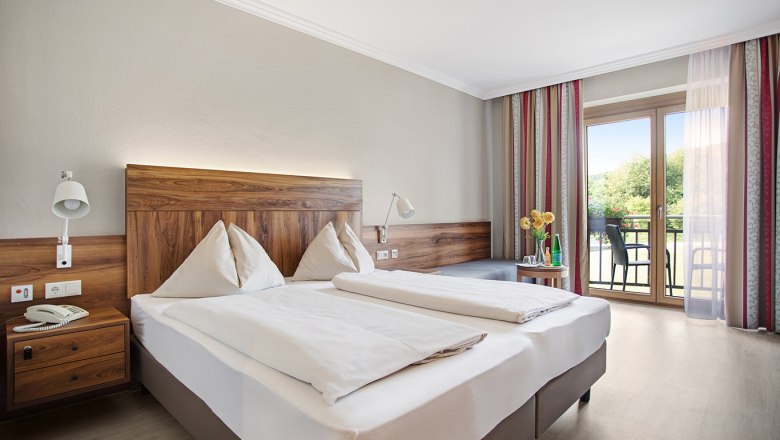 Ein gem&uuml;tliches Hotelzimmer mit Doppelbett, Holzm&ouml;beln und Balkon mit Blick ins Gr&uuml;ne.