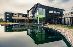 Modernes Wellnesshotel mit Holzfassade und gro&szlig;em Teich im Vordergrund.