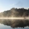 Ein Boot auf einem nebligen See vor einem bewaldeten Ufer bei Sonnenaufgang.