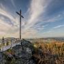 Ein Gipfelkreuz auf einem Felsen mit einer Aussichtsplattform, umgeben von Wald und einem weiten Blick &uuml;ber die Landschaft unter einem blauen Himmel.