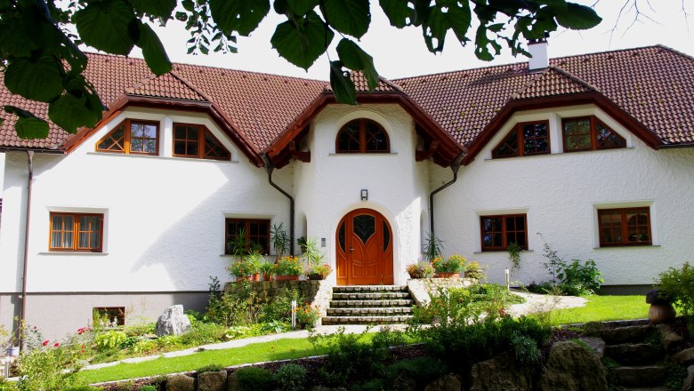 Ein gro&szlig;es, wei&szlig;es Haus mit rotem Ziegeldach und gepflegtem Garten, umgeben von B&auml;umen.