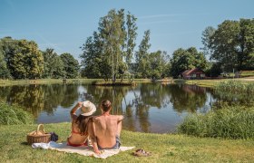Ein Paar sitzt auf einer Decke am Ufer eines Sees, umgeben von B&auml;umen und einem roten Geb&auml;ude im Hintergrund.