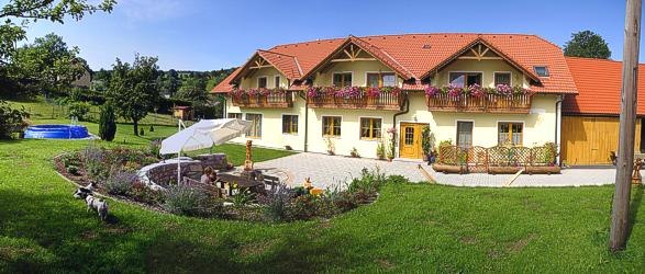Ein gro&szlig;es gelbes Haus mit roten D&auml;chern und Blumenk&auml;sten, umgeben von einem gepflegten Garten mit Sitzbereich und Sonnenschirm.
