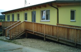 Gelbes Geb&auml;ude mit Holzrampe und Fenstern.