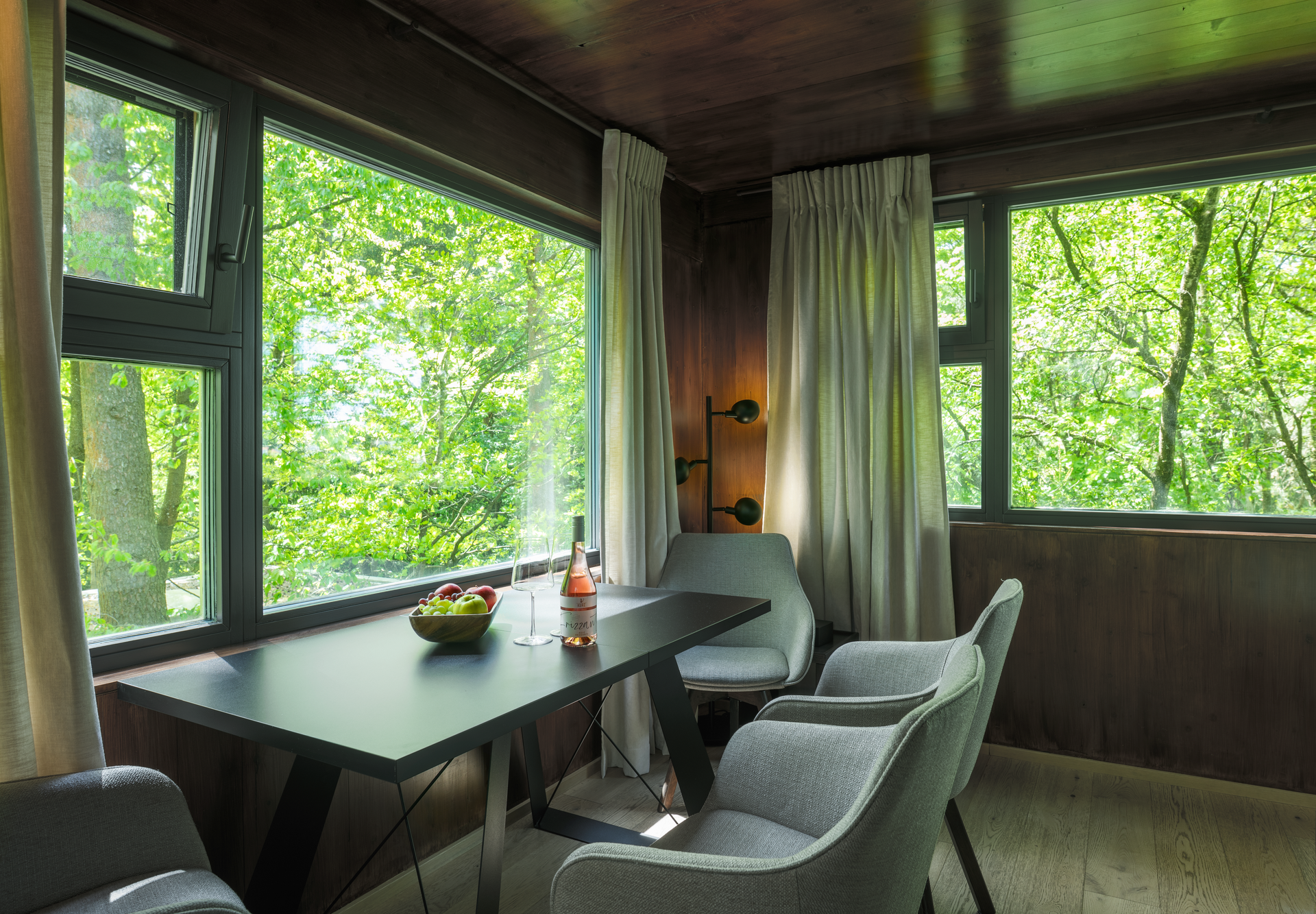 Innenansicht eines modernen Baumhauses mit Tisch, Stühlen und großen Fenstern mit Blick ins Grüne.