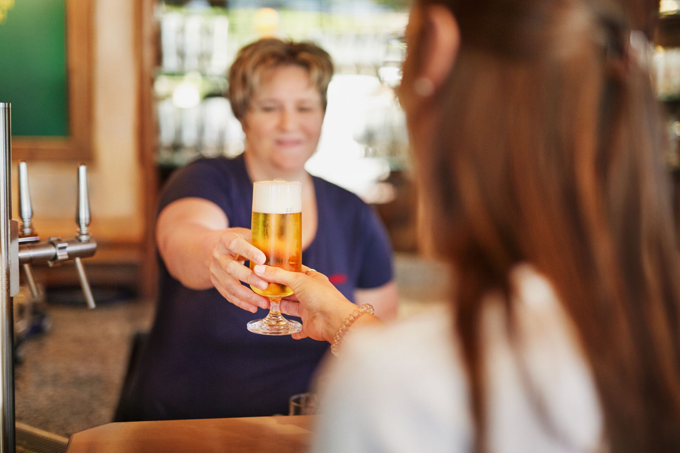 Eine Frau reicht einem Gast ein frisch gezapftes Bier in einem Glas über die Theke.