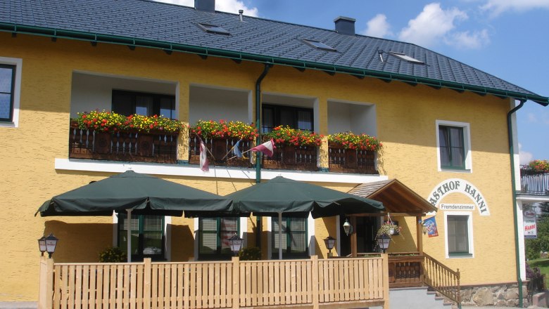 Gelbes Geb&auml;ude mit Balkon, Blumen und Terrasse mit Sonnenschirmen.