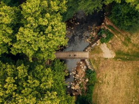 Zwei Personen kommen aus einem Waldst&uuml;ck und gehen &uuml;ber eine Br&uuml;cke zu einer Wiese