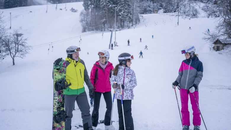 Gruppe von Skifahrern und Snowboardern im Schnee vor einer Skipiste mit Lift.