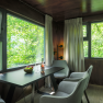 Innenansicht eines modernen Baumhauses mit Tisch, St&uuml;hlen und gro&szlig;en Fenstern mit Blick ins Gr&uuml;ne.