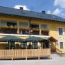 Gelbes Geb&auml;ude mit Balkon, Blumen und Terrasse mit Sonnenschirmen.