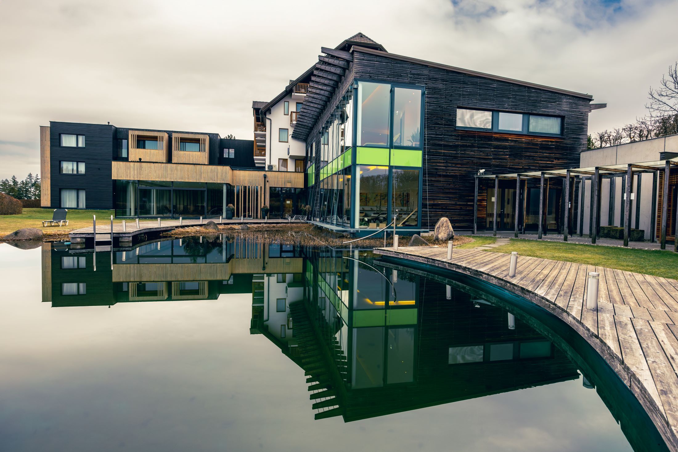 Modernes Wellnesshotel mit Holzfassade und großem Teich im Vordergrund.
