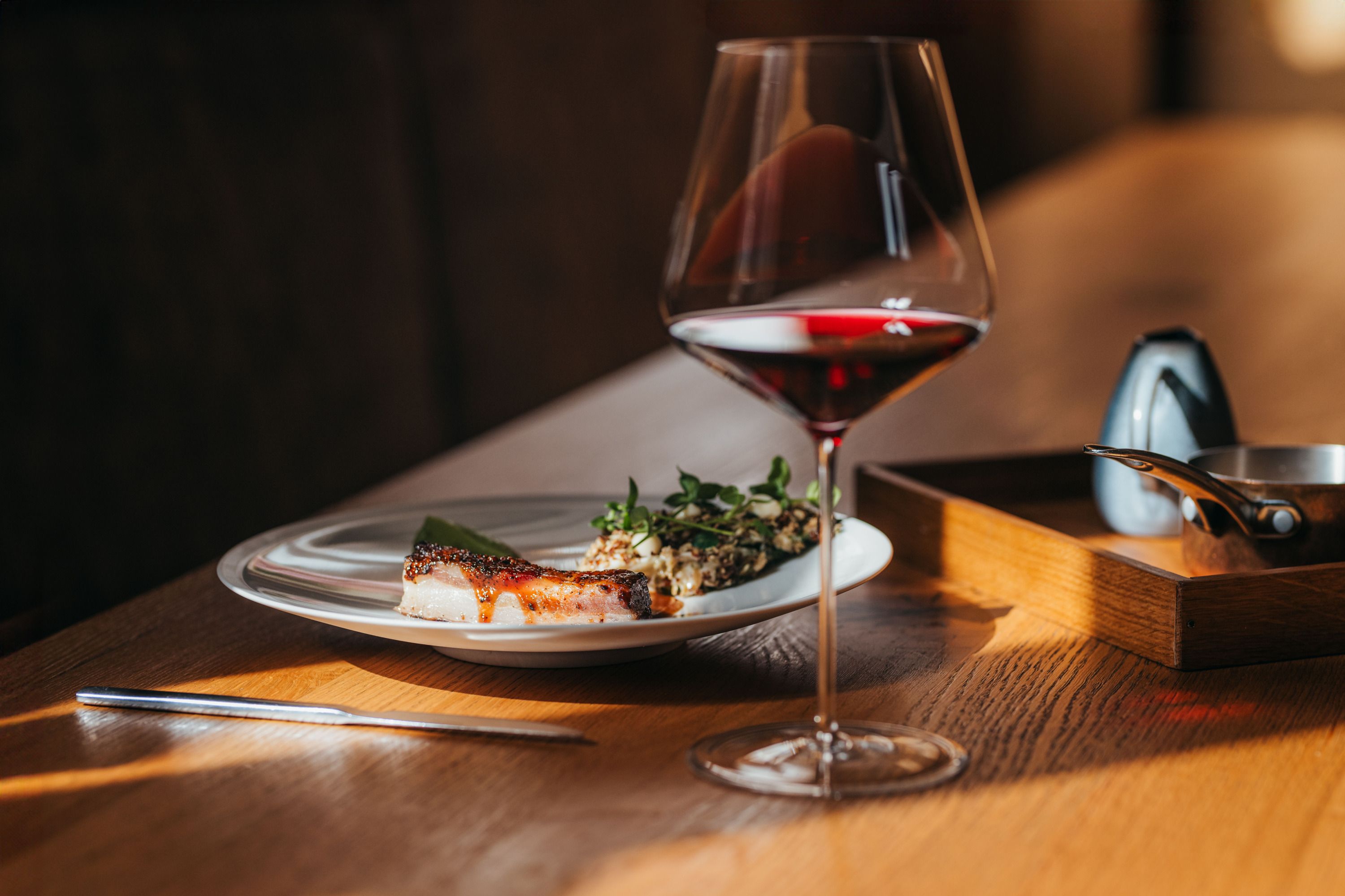 Ein Teller mit Lammbauch, Kopfsalat und Wasabicrème neben einem Glas Rotwein auf einem Holztisch.