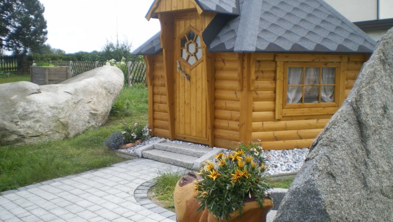 Grillhütte, © Grill Manfred Eine kleine, hölzerne Grillhütte mit grauem Dach, umgeben von Steinen und Pflanzen.