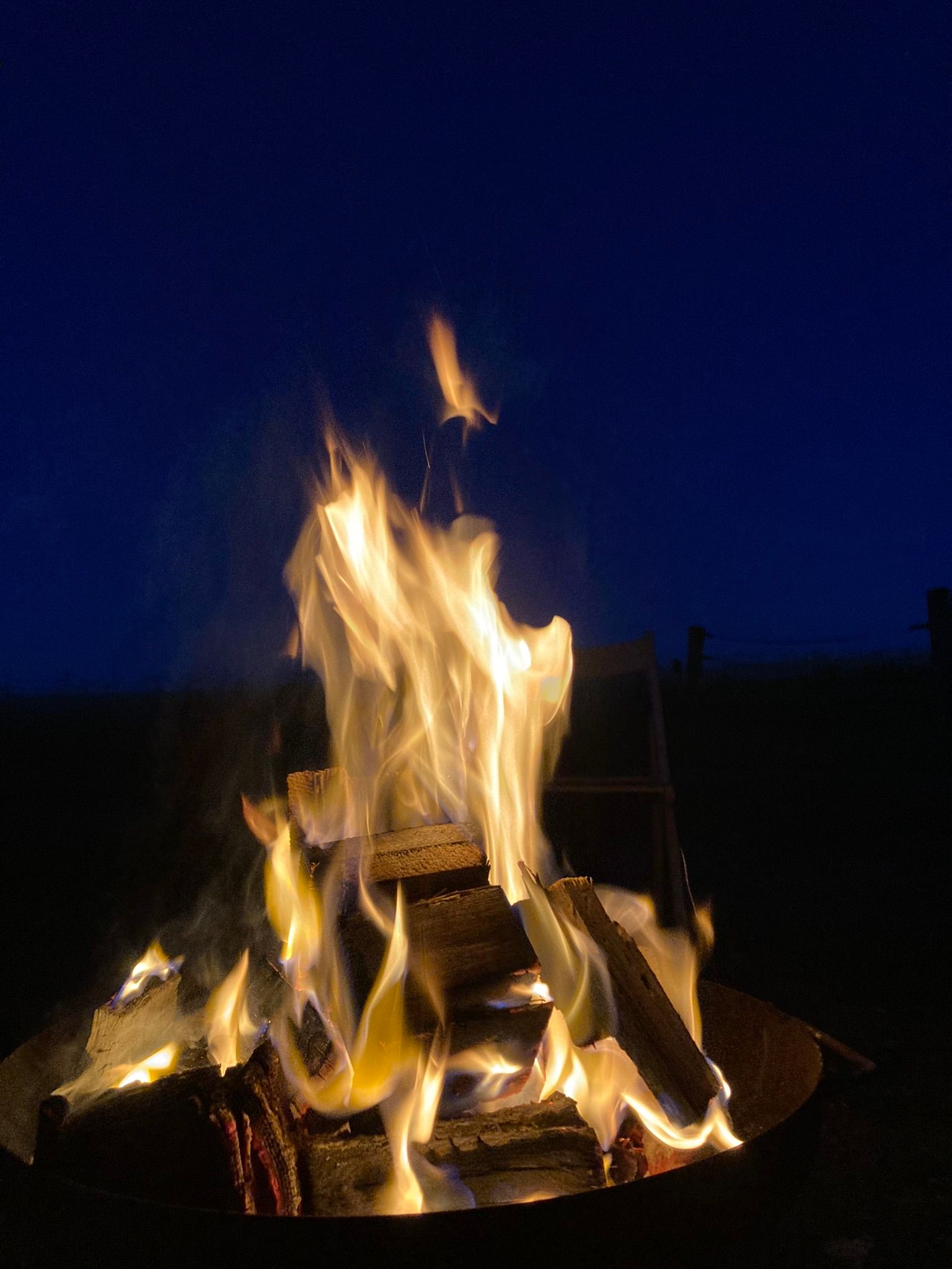 Ein Lagerfeuer brennt in der Dunkelheit, umgeben von Holzscheiten und Flammen, die in die Nacht aufsteigen.