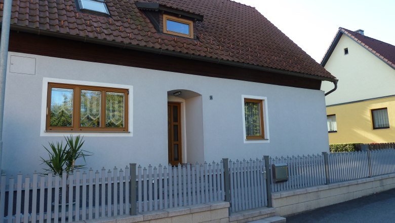 Sauschlager, © Sauschlager Ein Einfamilienhaus mit grauer Fassade, braunem Dach und Holztüren. Ein kleiner Zaun umgibt das Grundstück.