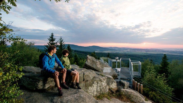 Zwei Personen sitzen auf Felsen bei der Aussichtsplattform Mandelstein und blicken in die Ferne. Der Himmel ist teils bewölkt, die Sonne geht unter.