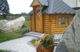 Eine kleine, h&ouml;lzerne Grillh&uuml;tte mit grauem Dach, umgeben von Steinen und Pflanzen.