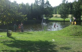 Menschen baden in einem Naturteich umgeben von B&auml;umen und Wiese.