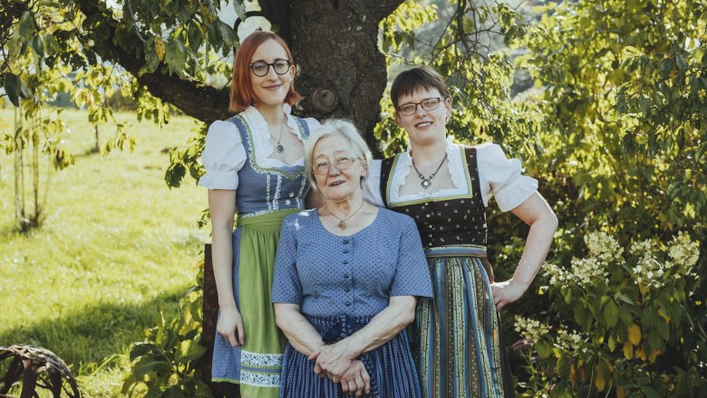 Drei Frauen in traditioneller Tracht stehen l&auml;chelnd unter einem Baum im Freien.