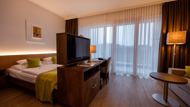 Modernes Hotelzimmer mit Doppelbett, Fernseher, Schreibtisch und gro&szlig;en Fenstern mit Vorh&auml;ngen.